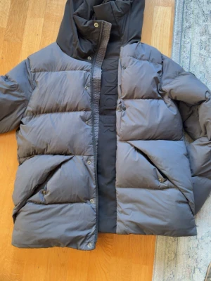Grå dunjacka från Moncler - Snygg grå dunjacka från Moncler med stor huva och breda, vadderade paneler. Jackan har tryckknappar, dragkedja och två stora fickor framtill. Moncler-logga på ärmen och diskret Moncler-tryck längs dragkedjan. Perfekt för kalla vinterdagar. RFID fungerar och qr koden fungerar helt.