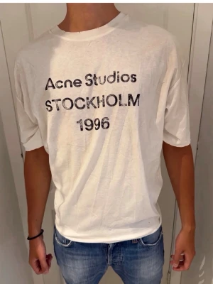 Vit Acne Studios t-shirt - Vit t-shirt från Acne Studios med tryck i svart text på bröstet där det står 'Acne Studios STOCKHOLM 1996'. T-shirten har en avslappnad passform och är gjord i mjuk bomull. Perfekt till jeans eller shorts.
