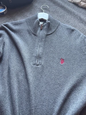 Grå stickad tröja från U.S. Polo Assn - Snygg grå stickad tröja från U.S. Polo Assn med halv dragkedja och ribbad hög krage. Tröjan har en liten röd logga broderad på bröstet och är perfekt för lager på lager. Materialet känns mjukt och bekvämt.
