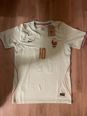 Frankrike fotbollströja Mbappé #10 - Snygg ljus fotbollströja med Frankrikes landslagsemblem och nummer 10 i guld. Mbappé står på ryggen. Tröjan har korta ärmar, tunna röda detaljer och randiga muddar i blått, vitt och rött. Perfekt för match eller träning.