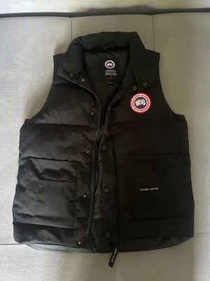 Äkta Canada Goose väst - Snygg svart dunväst från Canada Goose med klassisk logga på bröstet. Västen har hög krage, dragkedja och tryckknappar framtill samt två stora fickor. Perfekt för lager-på-lager och håller dig varm under kyliga dagar.