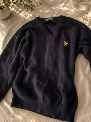 Lyle & scott v-ringad tröja - Mörkblå stickad tröja från Lyle & Scott med v-ringning och gul logga broderad på bröstet. Tröjan har långa ärmar och ribbade muddar vid ärmslut och nederkant. Perfekt för lager-på-lager och stilren look.