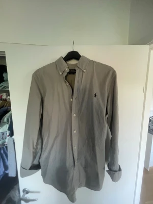 Beige/grön skjorta från Polo Ralph Lauren - Stilren beige skjorta från Polo Ralph Lauren i slim fit-modell. Skjortan har klassisk krage med knappar, långa ärmar och den ikoniska broderade loggan på bröstet. Perfekt för en clean och snygg look.