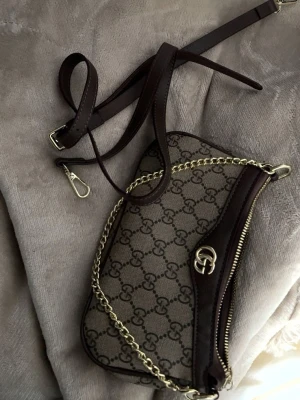 G Väska - Snygg beige handväska mörkbruna läderdetaljer. Väskan har guldfärgad kedja och avtagbar axelrem, samt dragkedja upptill. Perfekt för dig som gillar lyxiga accessoarer med ikonisk design. Helt ny