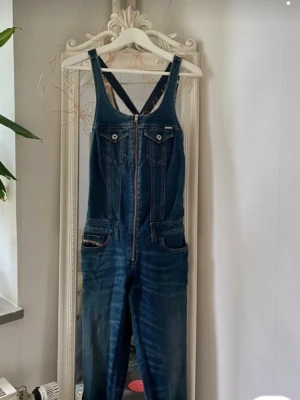 Blå denim jumpsuit med dragkedja - Snygg blå denim jumpsuit med dragkedja framtill, bröstfickor med knappar och fickor vid höfterna. Smala ben med dragkedjor vid bensluten ger en cool look. Tillverkad i stretchigt jeansmaterial för skön passform.