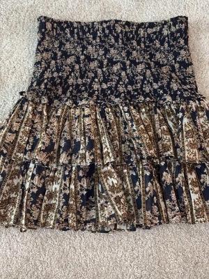 Blommig volangkjol Isabel Marant - Supersöt kort kjol från Isabel Étoile Marant med blommigt mönster i mörkblått, beige och brunt. Kjolen har smock i midjan och flera volanger som ger en flowy känsla. Perfekt för dig som gillar boho-vibbar och vill ha något unikt i garderoben.