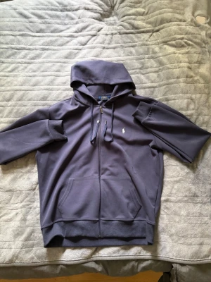 Mörkblå zip hoodie från Polo Ralph Lauren - Mörkblå hoodie med dragkedja från Polo Ralph Lauren. Klassisk design med huva och dragsko, två fickor framtill och den ikoniska vita logon broderad på bröstet. Tillverkad i mjukt sweatshirtmaterial, perfekt för en avslappnad stil.