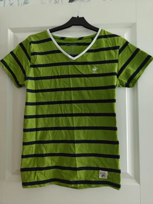 Randig grön t-shirt från Beverly Hills Polo Club - Grön t-shirt med mörkblå horisontella ränder från Beverly Hills Polo Club. T-shirten har vit v-ringad krage och korta ärmar. Tillverkad i mjuk bomull och har en liten logga broderad på bröstet.