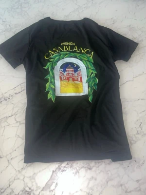 Svart t-shirt med Casablanca-tryck - Säljer en svart t-shirt med färgglatt tryck på både bröstet och ryggen. Motivet visar en byggnad med texten 'Avenida Casablanca' och gröna blad runt fönstret. T-shirten har rund hals och korta ärmar, perfekt för en chill och stilren look.