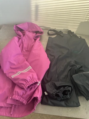 Rosa regnjacka från Kaxs.. - Svart jumpsuit med hängslen och elastiska benslut. Tillverkad i vattenavvisande material, perfekt för utomhusbruk. Justerbara axelband för skön passform och reflexdetaljer för extra synlighet. Enkel att dra på över andra kläder.