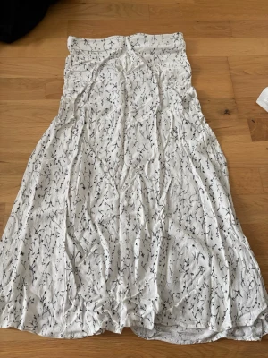Vit kjol med blommönster - Långkjol i vit färg med svart blommönster. Kjolen har en rak och luftig passform och perfekt för varma dagar. Tillverkad i ett mjukt och lätt tyg som känns skönt mot huden. Lite genomskinlig men inget som inte går att täcka med cykelbyxor under 