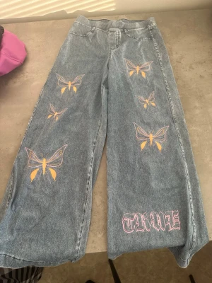 Blå jeans med fjärilar från H&M. - Säljer ett par blå raka jeans från H&M med färgglada fjärilsbroderier och trycket 'TRUE' i vitt och rosa på ena benet. Jeansen har hög midja och avslappnad passform, perfekta för en trendig och personlig stil.
