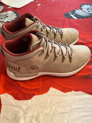 Beige Timberland kängor i mocka - Snygga beige kängor från Timberland i mocka med vita sulor och röda detaljer på insidan. Klassisk Timberland-logga på sidan och grova snören för en sportig look. Perfekta för dig som vill ha både stil och komfort i vardagen.
