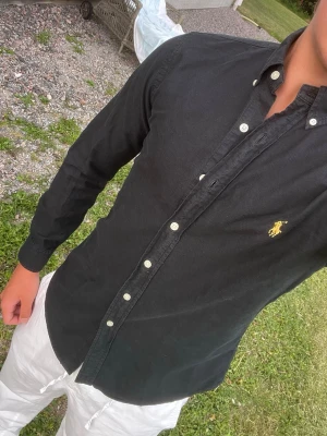 Svart skjorta från Polo Ralph Lauren - Säljer en svart långärmad skjorta från Polo Ralph Lauren med klassisk button-down krage och gul broderad logga på bröstet. Skjortan har vita knappar och är i ett mjukt bomullsmaterial. Perfekt till jeans eller chinos!