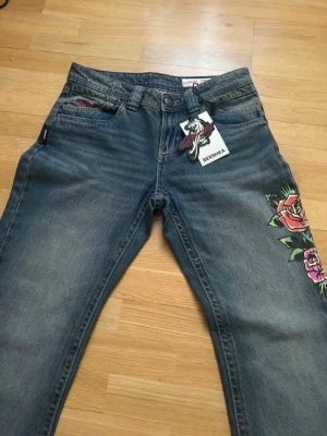 Blå jeans från Bershka x ed Hardy  - Snygga blå jeans från Bershka x ed Hardy  med färgglada rosor längs ena benet. Jeansen har strass, låg midja och bootcut. Materialet är denim i bomull och de har en unik detalj med strass text på bakfickan.
