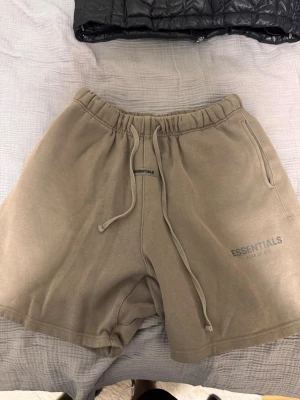 Beige shorts från Essentials Fear of God - Snygga beige shorts från Essentials Fear of God med elastisk midja och snörning. Mjukt material och avslappnad passform, perfekt för chill dagar. Diskret logga på benet och klassisk design som funkar till det mesta.