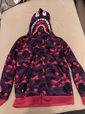 Lila och rosa camo hoodie med hajmotiv - Säljer en hoodie med dragkedja i lila och rosa camomönster. Huvan har ett hajansikte med tänder och ögon tryckt på, vilket ger en unik streetwear-look. Hoodien har fickor framtill och ribbade muddar vid ärmar och nederkant. Perfekt för dig som vill sticka ut.