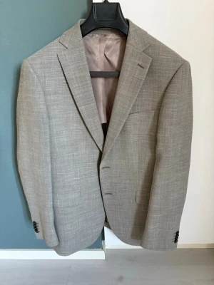 Beige kavaj från Long Island - Stilren beige kavaj från Long Island i storlek 52. Kavajen har två knappar framtill, klassisk krage och diskreta fickor. Materialet är en blandning av linne och viskos, vilket ger en lätt och luftig känsla. Perfekt för en dressad look.