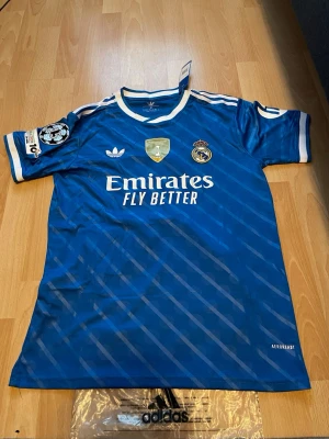 Blå Real Madrid matchtröja Adidas - Snygg blå Real Madrid matchtröja från Adidas med vita detaljer och tryck. Tröjan har klubbmärke, sponsorlogga och Champions League-märke på ärmen. På ryggen står det Valverde och nummer 8. Tillverkad i lätt och ventilerande material.