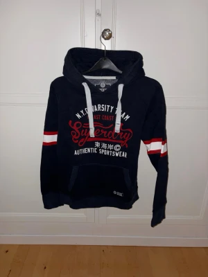 Superdry Hoodie - Snygg mörkblå hoodie med vit och röd text på bröstet och vita samt röda ränder på ärmarna. Stora vita dragsnören i huvan och känguruficka framtill. Perfekt för en sportig och avslappnad stil.