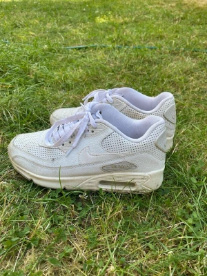 Vita Nike Air Max sneakers - Säljer ett par helvita Nike Air Max sneakers med perforerad ovandel och klassisk synlig luftdämpning i sulan. Skorna har låga skaft, vita snören och diskret Nike-logga på sidan. Perfekta för dig som gillar stilrena och sportiga sneakers.