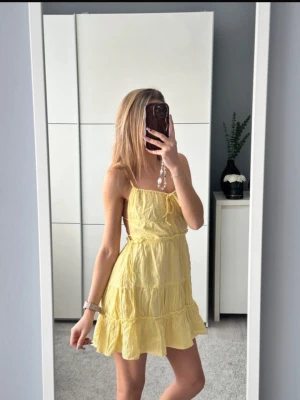Gul kort sommarklänning med volanger - Supersöt gul kort klänning med tunna axelband och volangdetaljer. Klänningen har öppen rygg med knyt i midjan och är gjord i ett lätt, luftigt material som passar perfekt till varma dagar.