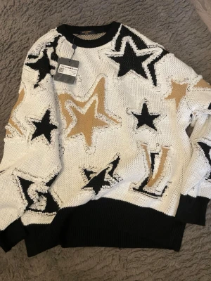 Stickad tröja med stjärnor - Cool stickad tröja i vitt med stora stjärnor i svart och beige. Tröjan har svarta muddar vid hals, ärmslut och nederkant. Oversized passform och trendig design med fransiga kanter runt stjärnorna. Perfekt för dig som vill sticka ut.