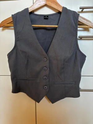 Mörkgrå väst med knappar storlek XS - Mörkgrå väst med fyra knappar framtill och två dekorativa fickor. Västen har djup v-ringning och figurnära passform. Materialet är polyester