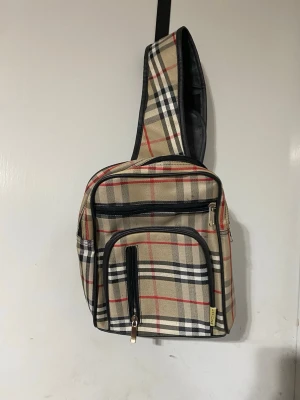 Rutig crossbody ryggsäck  - Snygg ryggsäck i beige med klassiskt rutigt mönster i svart, vitt och rött. Väskan har flera fack med dragkedja och en bred axelrem i svart. Perfekt för dig som vill ha en trendig och praktisk väska till vardagen. Helt ny 