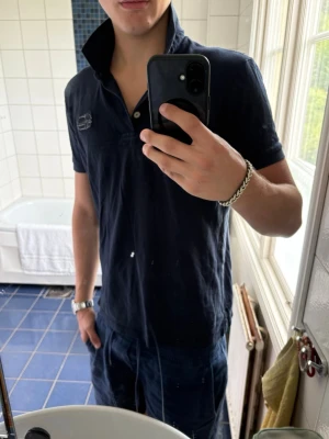 Mörkblå napapijri piké med krage - Snygg mörkblå pikétröja med klassisk krage och korta ärmar. Tröjan har knappar vid halsen och en diskret patch på bröstet. Perfekt till jeans eller shorts för en avslappnad stil.