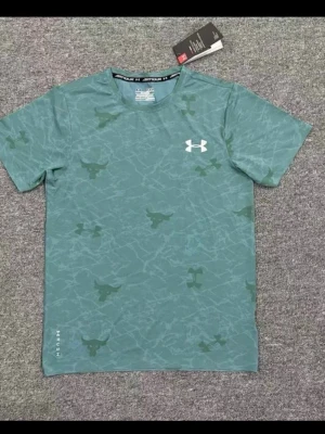 Grön mönstrad t-shirt Under Armour - Snygg grön t-shirt från Under Armour med diskret mönster och logga på bröstet. T-shirten har korta ärmar och rund halsringning. Perfekt för dig som gillar sportig stil och vill ha något bekvämt till vardags.