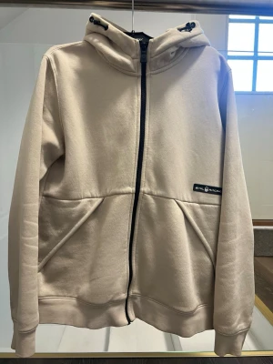 Beige sail racing hoodie med dragkedja och huva - Stilren beige hoodie från sail racing med hel dragkedja framtill, justerbar huva med snörning och två stora fickor. Liten svart logotyp på bröstet. Mjuk och skön bomullsblandning, perfekt för en avslappnad look.