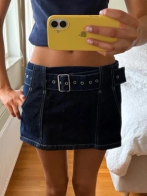 Mörkblå jeanskjol med bälte - Snygg mörkblå jeanskjol i kort modell med markerade vita sömmar och ett brett bälte med silverfärgat spänne. Kjolen har fickor på sidorna och dragkedja i sidan