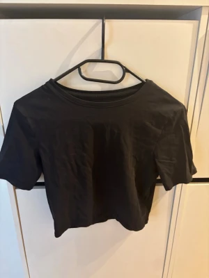 Svart croppad t-shirt - En enkel svart croppad t-shirt med rund hals och korta ärmar. Perfekt basplagg som passar till det mesta och är lätt att styla. Tillverkad i mjuk bomull för en skön känsla.