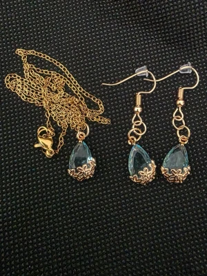 Set med halsband och örhängen i guld - Snyggt smyckeset med halsband och matchande örhängen i guldfärgad metall. Båda har droppformade ljusblå stenar infattade i dekorativa guldfärgade detaljer. Perfekt för att lyfta din outfit med lite extra bling.