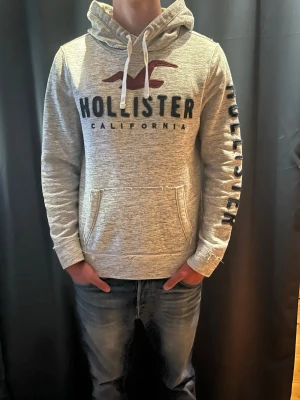 Hollister hoodie  - Snygg gråmelerad hoodie från Hollister med stor logga. Har en stor huva med vita snören och känguruficka framtill. Hollister-tryck även på ena ärmen. Mjuk insida och den är aldrig använd och bär storleken M 🤝