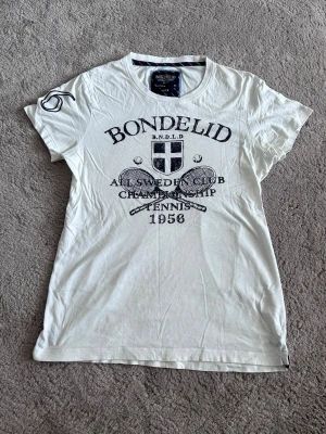 Vit t-shirt från Bondelid med tryck - Vit t-shirt från Bondelid med stort svart tryck framtill och broderad siffra '56' på ena ärmen. T-shirten har rund halsringning och klassisk passform. Perfekt för dig som gillar sportig stil och tennisinspirerade detaljer.