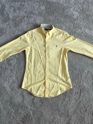 Gul skjorta från Ralph Lauren - Säljer en ljusgul skjorta från Ralph Lauren med klassisk button-down krage och broderad logga på bröstet. Skjortan har långa ärmar och vita knappar framtill. Perfekt för dig som gillar stilrena och fräscha plagg. ALDRIG ANVÄND!