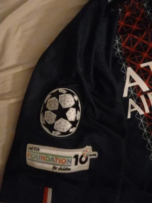 PSG matchtröja Champions League Nike - Säljer en mörkblå PSG matchtröja från Nike med Champions League-märke på ärmen och Qatar Airways som sponsor på bröstet. Tröjan har röd och vit grafisk detalj i mitten och PSG-logga på bröstet. Perfekt för fotbollsfans!