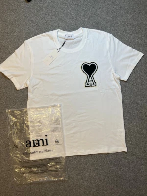 Vit t-shirt från Ami med svart hjärtdetalj - Vit t-shirt från Ami Alexandre Mattiussi med en stor svart hjärtdetalj och bokstaven A på bröstet. Klassisk passform med rund hals och korta ärmar. Perfekt för en stilren och trendig look.