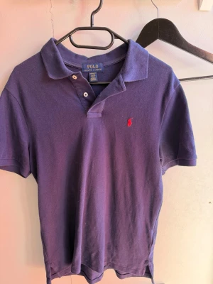 Lila pikétröja från Polo Ralph Lauren - Klassisk lila pikétröja från Polo Ralph Lauren i custom fit-modell. Tröjan har korta ärmar, krage och två knappar vid halsen. Ikoniska röda logotypen broderad på bröstet. Tillverkad i bomull för en bekväm känsla.