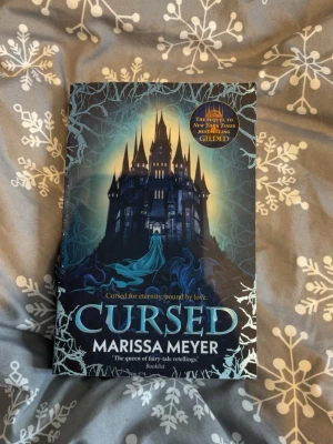Cursed av Marissa Meyer - Säljer boken 'Cursed' av Marissa Meyer, en fantasyroman med ett mörkt slott på omslaget och blåa, svarta och silvriga toner. Boken är på engelska och är en uppföljare till 'Gilded'. Perfekt för dig som gillar sagoinspirerade berättelser och magiska världar.
