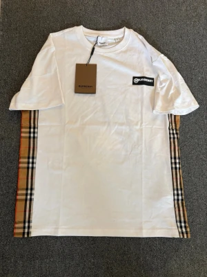 Unik vit T shirt  - Vite t-shirt från Burberry med klassiska rutiga paneler längs sidorna och en svart Burberry-logga på bröstet. T-shirten har rund hals och korta ärmar. Tillverkad i mjuk bomull för en skön känsla och stilren look.