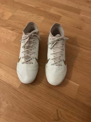 Vita Nike fotbollsskor med guldlogga - Säljer ett par vita Nike fotbollsskor med guldiga swoosh-loggor på sidorna. Skorna har snörning framtill och sulan är svart med orange dobbar, perfekt för spel på gräs. Ovandelen är i syntetmaterial för bra bollkontroll och lätt känsla.