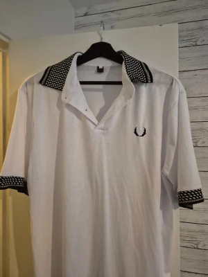 Vit piké med svartvitt mönstrad krage - Vit pikétröja från Fred Perry med svartvitt mönstrad krage och ärmslut. Klassisk logga broderad på bröstet. Tröjan har korta ärmar och normal passform, perfekt för en clean och stilren look.