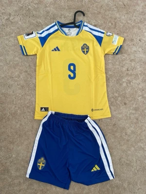 Sverige fotbollströja och shorts Adidas - Säljer ett matchställ från svenska landslaget med gul tröja och blå shorts. Tröjan har nummer 9 och ISAK på ryggen, samt landslagets emblem och Adidas-logga. Shortsen är blå med vita ränder och har också landslagets märke. Perfekt för fotbollsträning eller match.