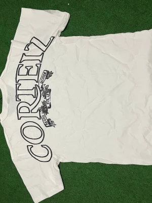 Vit Corteiz t-shirt med tryck - Vit t-shirt från Corteiz med stort svart tryck på ryggen och mindre logga framtill. Oversized passform och klassisk rund hals. Tillverkad i mjuk bomull, perfekt för en avslappnad streetwear-look.
