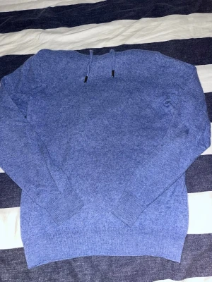 Blå merionoulls hoodie  - Den första bilden är denna färgen tröjan kommer vara. Mysig mörkblå hoodie med huva. Tillverkad i ett mjukt, stickat material som känns skönt mot huden. Långa ärmar och ribbade muddar vid ärmslut och nederkant ger en clean look. Perfekt för dig som gillar enkel och stilren design.