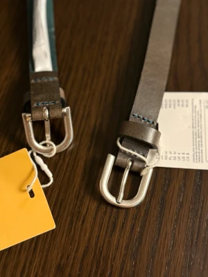 Brunt skärp i skinn med silverfärgat spänne - Snyggt brunt bälte i skinn med robust silverfärgat metallspänne och diskreta blå sömmar vid spännet. Perfekt accessoar för att lyfta din outfit och ge en stilren touch. Passar till både jeans och byxor.