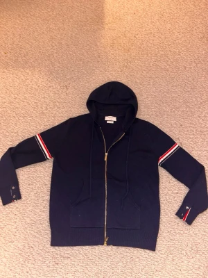 Mörkblå hoodie från Webolion - Snygg mörkblå hoodie från tom browne med dragkedja och huva. Ärmarna har röda och vita ränder samt knappar vid mudden. Tillverkad i Italien och har två stora fickor framtill. Perfekt för en avslappnad och trendig stil.